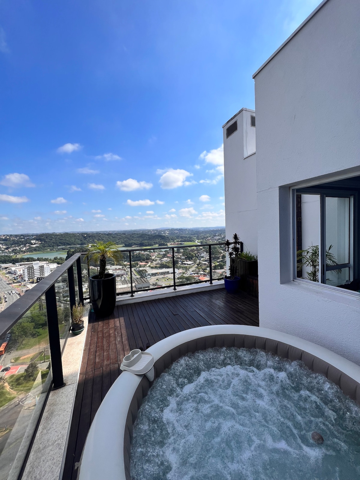 A spacious outdoor balcony features a jacuzzi overlooking the cityscape. The area is surrounded by glass railings, allowing unobstructed views. Potted plants add greenery, while the bright blue sky is dotted with fluffy clouds.