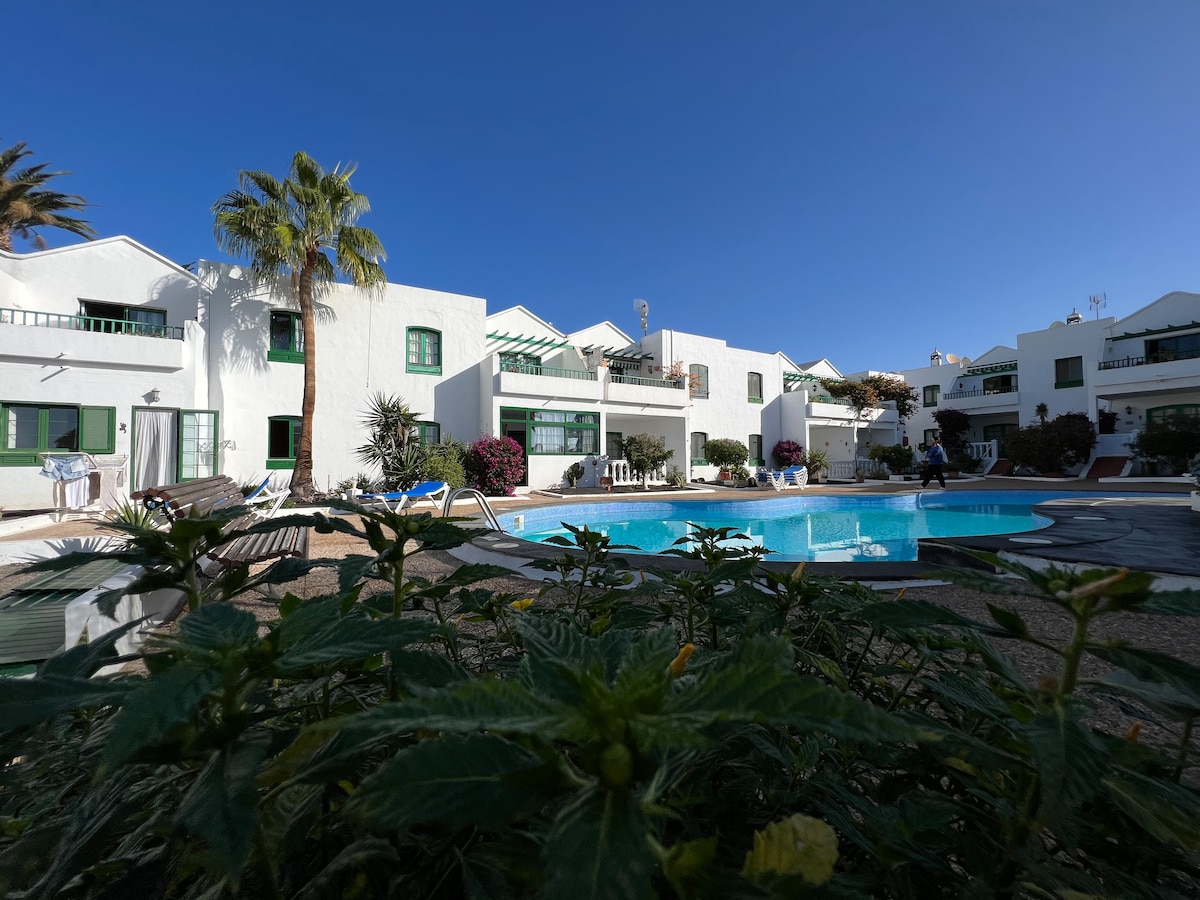 A tranquil pool area is surrounded by white buildings, featuring greenery and palm trees. Sun loungers are arranged around the pool's edge, offering a serene environment. The clear blue sky enhances the inviting atmosphere of the complex.