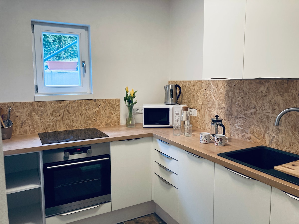 A modern kitchen is equipped with essential appliances including an oven, microwave, and kettle. Natural light enters through a small window, highlighting a countertop with a sink and coffee maker. Decorative flowers are placed in a vase for a touch of color.