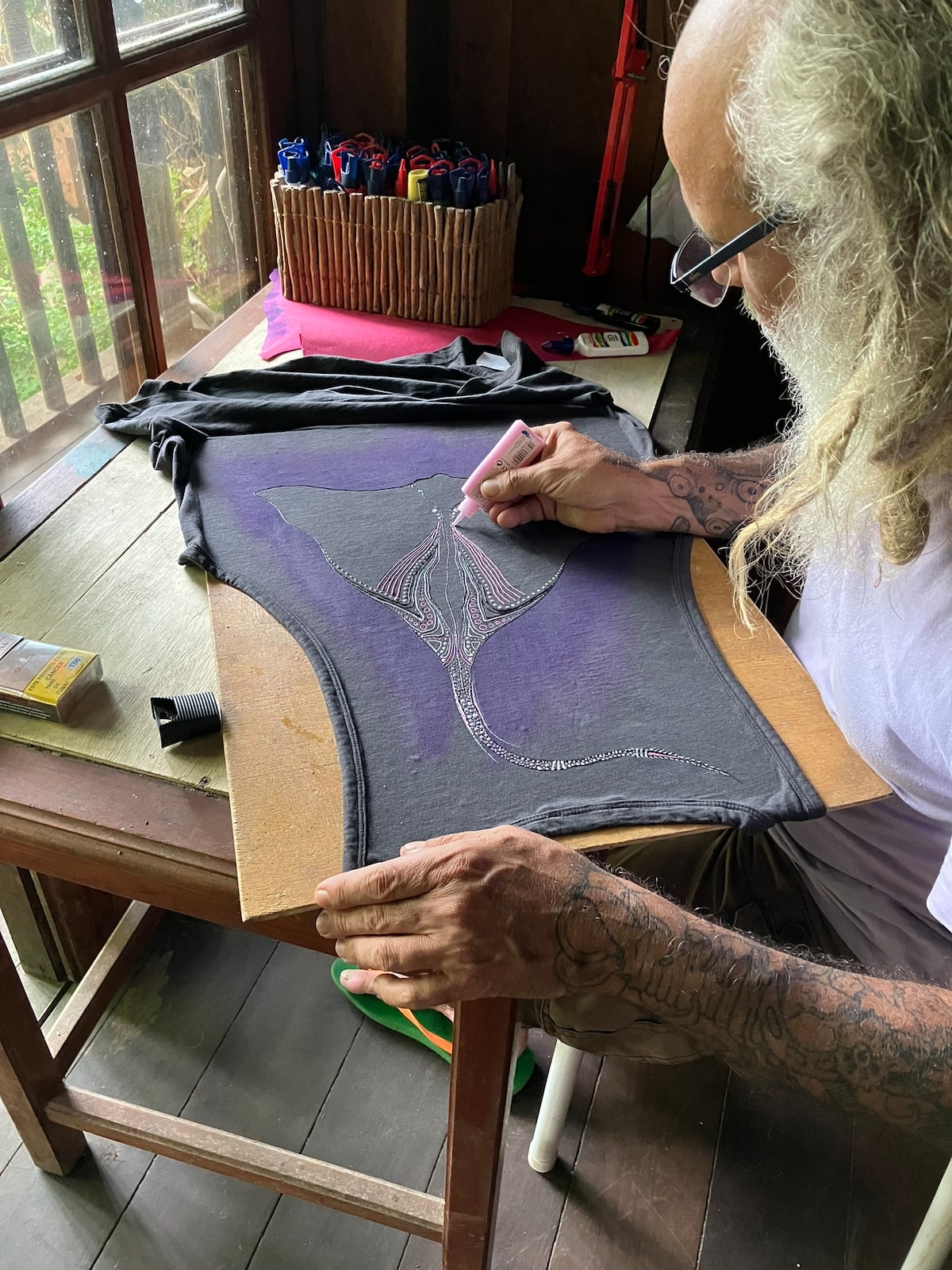 An individual is seen creating art on a fabric piece at a wooden table. A variety of tools and materials are visible on the table's surface. Natural light floods the space through large windows, highlighting the artistic process. The surrounding area features wooden beams and a lush view outdoors.
