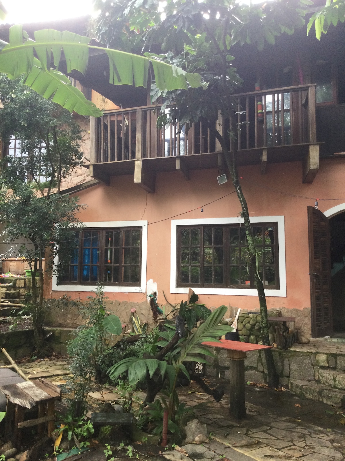 A two-story house is framed by lush greenery and large trees. The ground floor features multiple rectangular windows, while a balcony extends from the upper level. A small outdoor table is surrounded by tropical plants, enhancing the inviting ambiance of the garden setting.