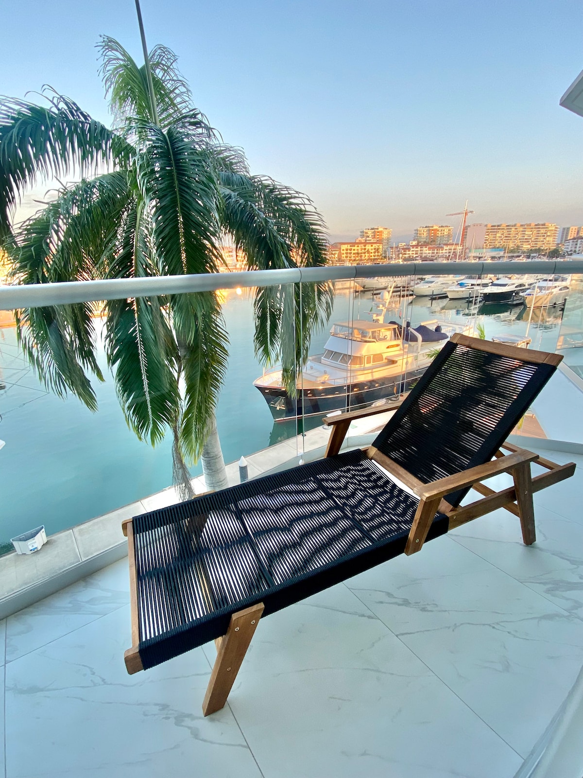 A sleek black lounge chair is positioned on a private balcony, which overlooks a marina filled with boats. Lush palm trees sway gently in the breeze, enhancing the serene waterfront setting. Soft sunlight reflects off the water, creating a calm and inviting atmosphere.