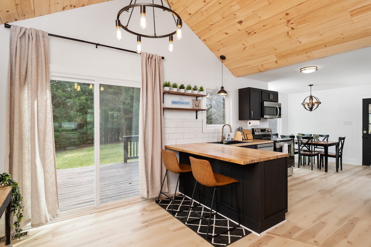 A modern kitchen features a wooden island with two stylish bar stools. Large windows allow natural light, offering views of the greenery outside. A dining area with a glass top table and black chairs complements the sleek cabinetry and stainless-steel appliances.