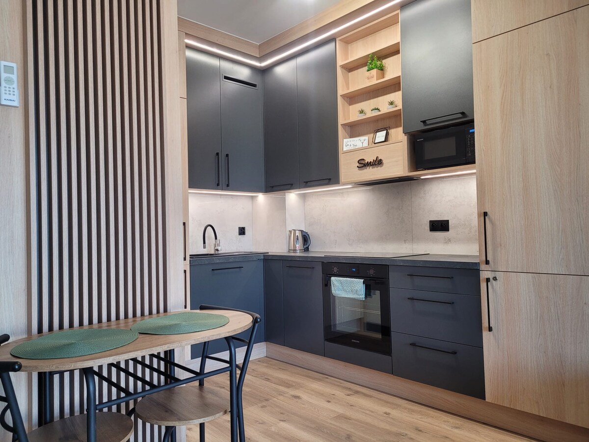 A modern kitchen features sleek black cabinetry and wooden accents, complemented by under-cabinet lighting. Equipped with a microwave and oven, it offers a functional design. A small dining table with two chairs rests nearby, enhancing the space's practicality.