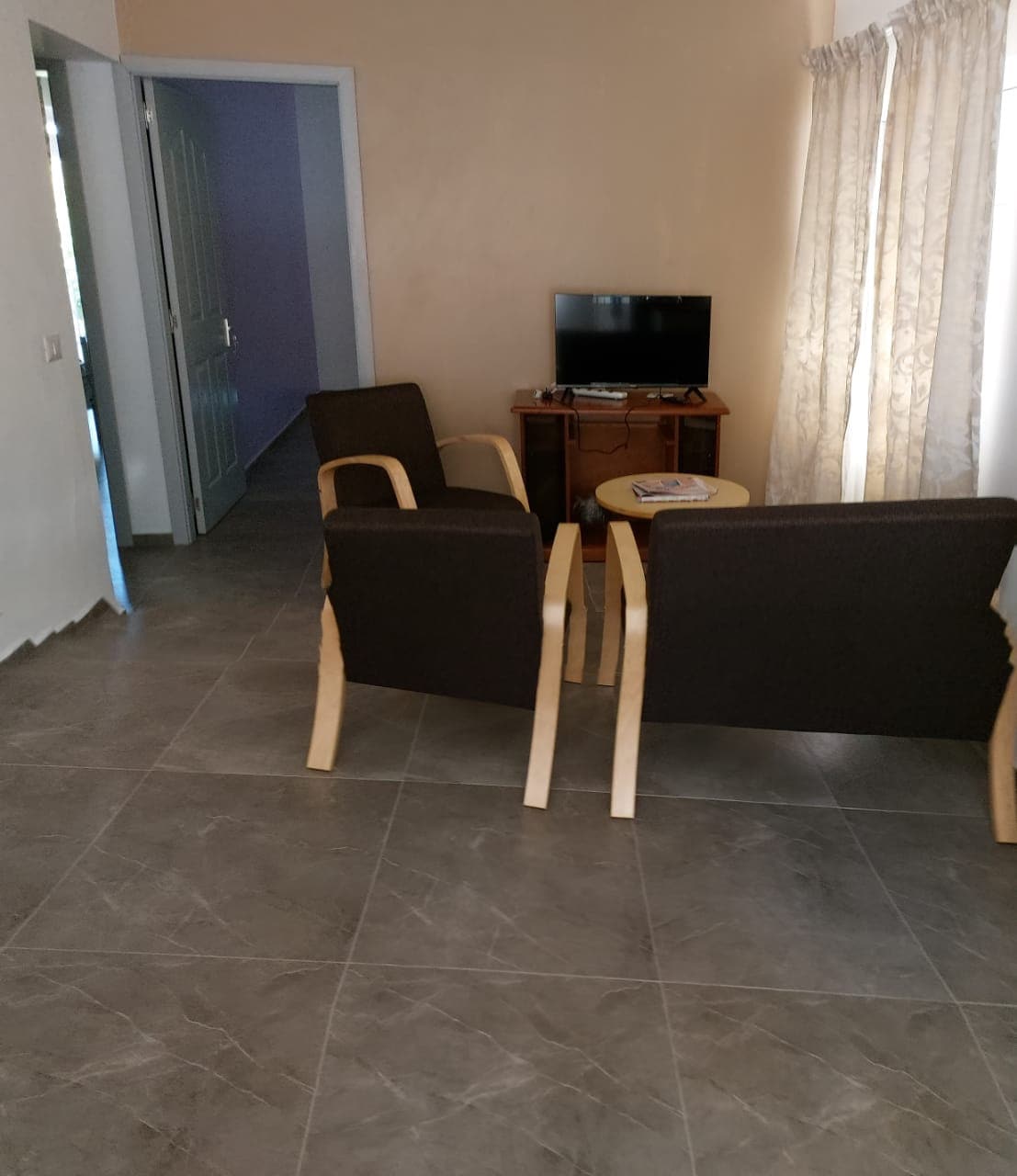 A cozy seating area features two wooden chairs facing a small round table, with a television on a wooden stand nearby. Light-colored curtains frame a window, allowing natural light to fill the space. The floor is covered in sleek gray tiles.