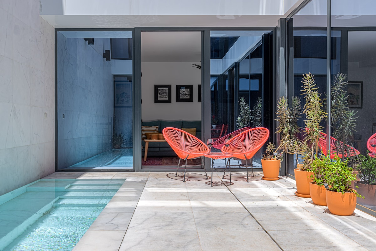 A serene outdoor space features two distinct red chairs surrounding a glass table. Potted plants are positioned along the marble terrace, and a private pool reflects the sunlight, enhancing the tranquil atmosphere of the area adjacent to the living space.
