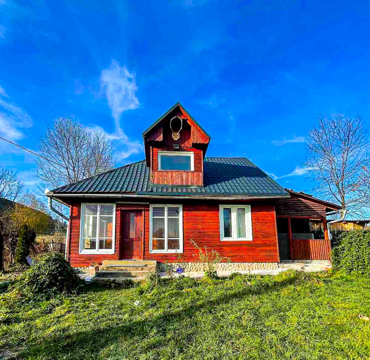 A vibrant red house is set against a bright blue sky, featuring multiple large windows that invite natural light. The sloped roof adds charm, while a grassy area surrounds the home, contributing to a welcoming exterior. Trees can be seen in the background.