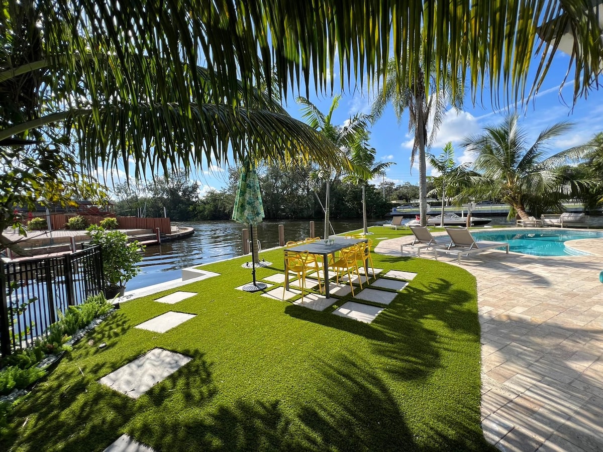 Lush greenery surrounds a spacious shared backyard featuring a well-kept pool. Two dining tables and lounge chairs are positioned on the manicured lawn, with a tranquil canal visible in the background. Sunlight filters through tall tropical palm trees, creating a serene outdoor setting.