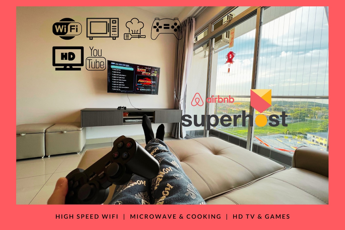A modern living area is depicted with a comfortable sectional sofa and a large screen TV displaying various entertainment options. A gaming controller sits in hand, indicating a space designed for leisure. Large windows offer a view of the surroundings, allowing natural light to fill the room.