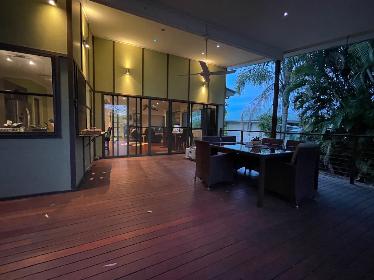 A spacious covered deck features a large dining table surrounded by wicker chairs, ideal for outdoor gatherings. Large sliding doors provide access to the interior living space, while ceiling fans above enhance airflow. Lush greenery surrounds the area, adding to its natural charm.