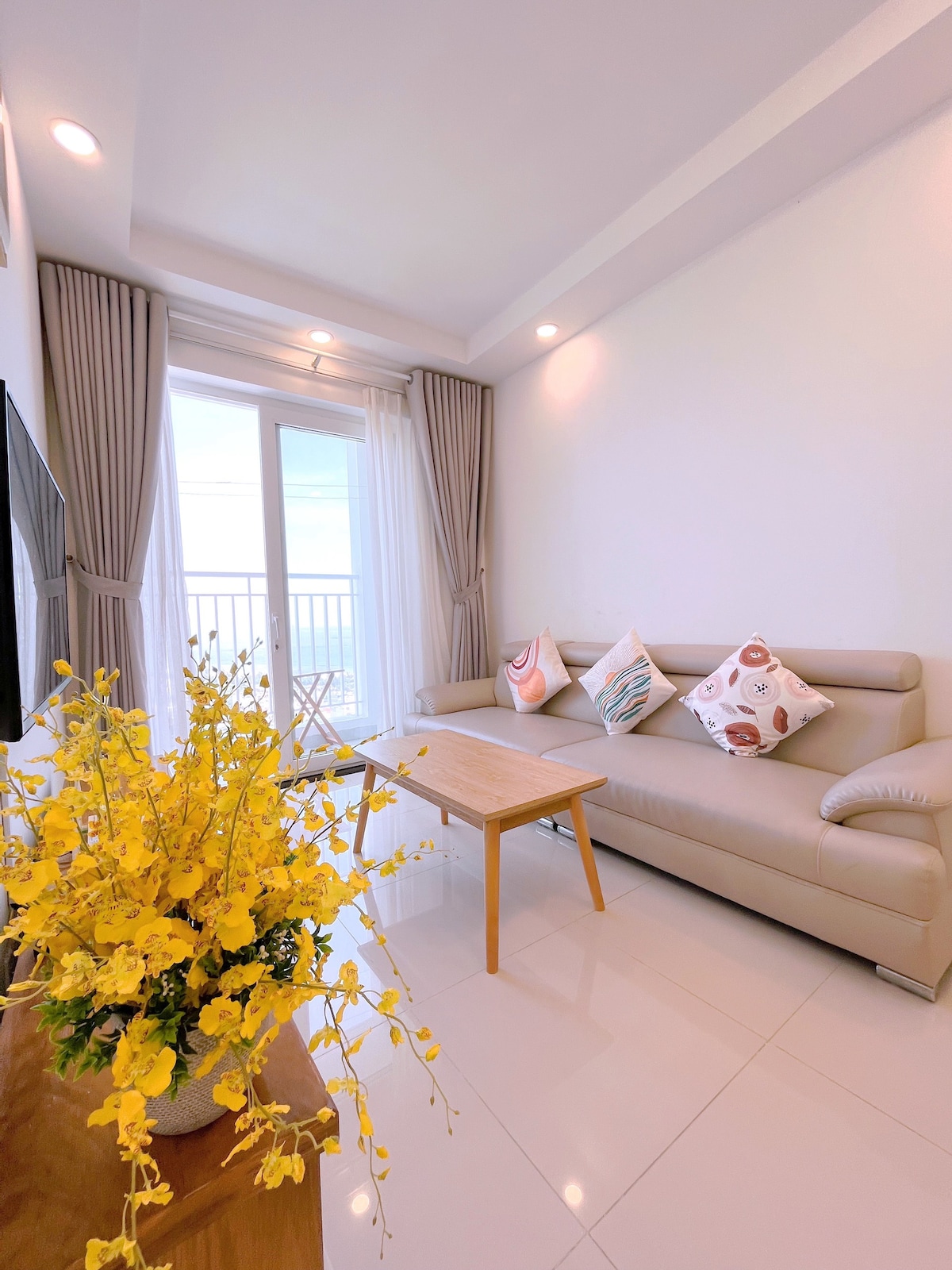 A well-lit living area features a light-colored sofa adorned with decorative pillows. A small wooden table occupies the center, complemented by a vibrant flower arrangement. Floor-to-ceiling curtains frame the balcony doors, allowing natural light to enhance the space.