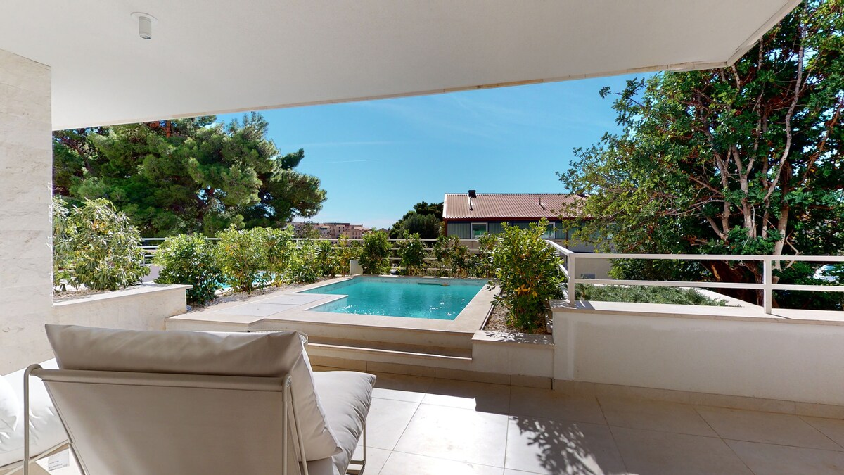 A spacious terrace features comfortable seating with a view of the private pool surrounded by lush greenery. Sunlight brightens the area, and a fence provides privacy while enhancing the natural surroundings.