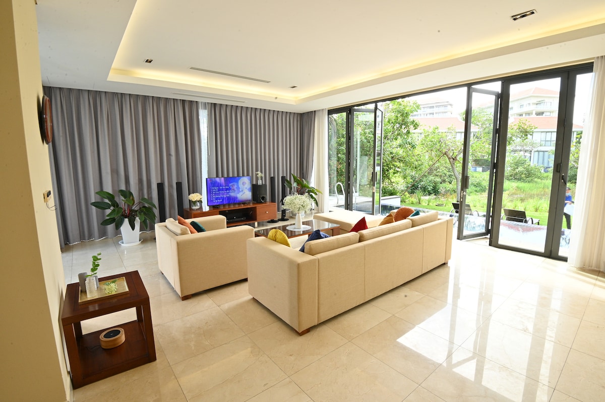 A spacious living area is highlighted by large glass doors, offering a view of the greenery outside. Soft seating arrangements with colorful cushions are positioned around a coffee table. Natural light illuminates the room, enhancing the serene ambiance.