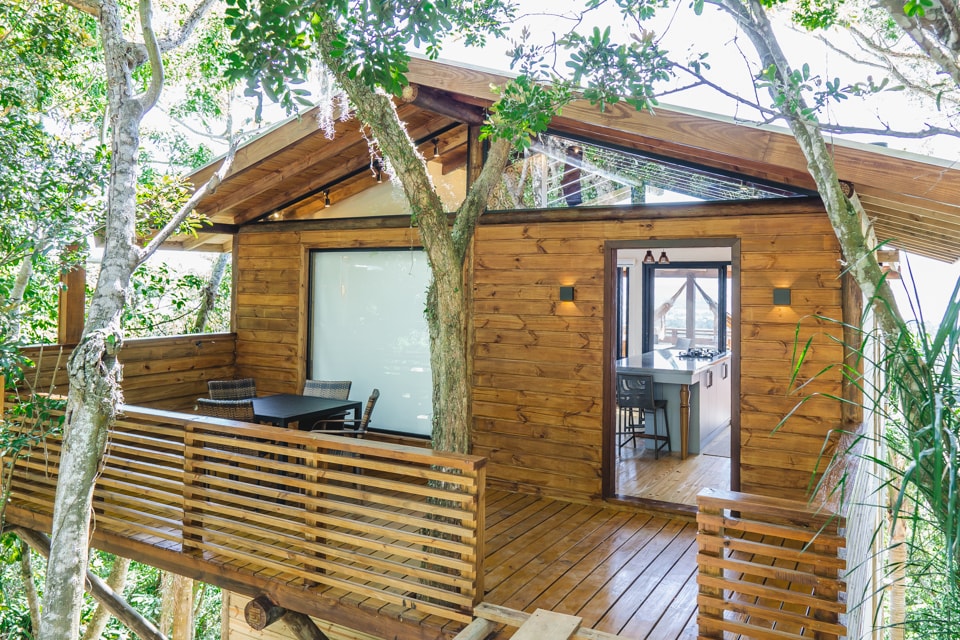 A contemporary rustic house is nestled among trees, featuring a wooden deck with table and chairs. Large windows invite natural light, while the doorway leads into an open space that connects to the kitchen. The exterior is enhanced by lush green foliage.