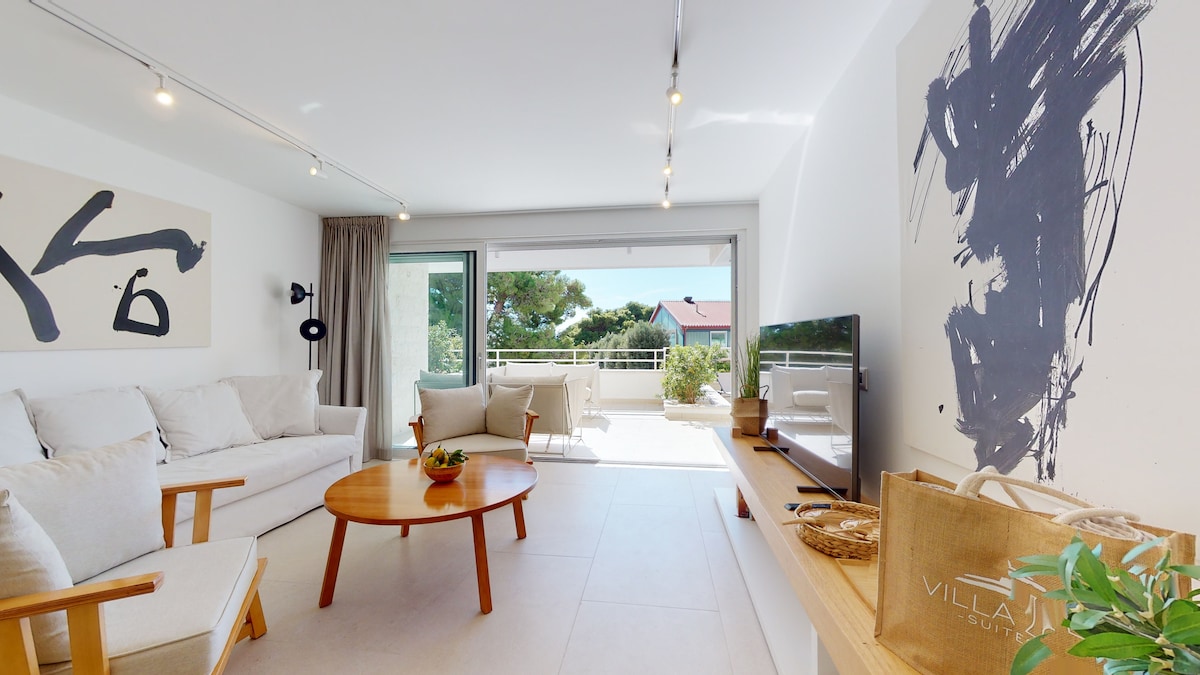 A modern living area features a comfortable sofa and a round wooden table. Natural light floods the space through large sliding glass doors, which open to a private terrace. A flat-screen television rests on a minimalist shelving unit, complementing the contemporary decor.