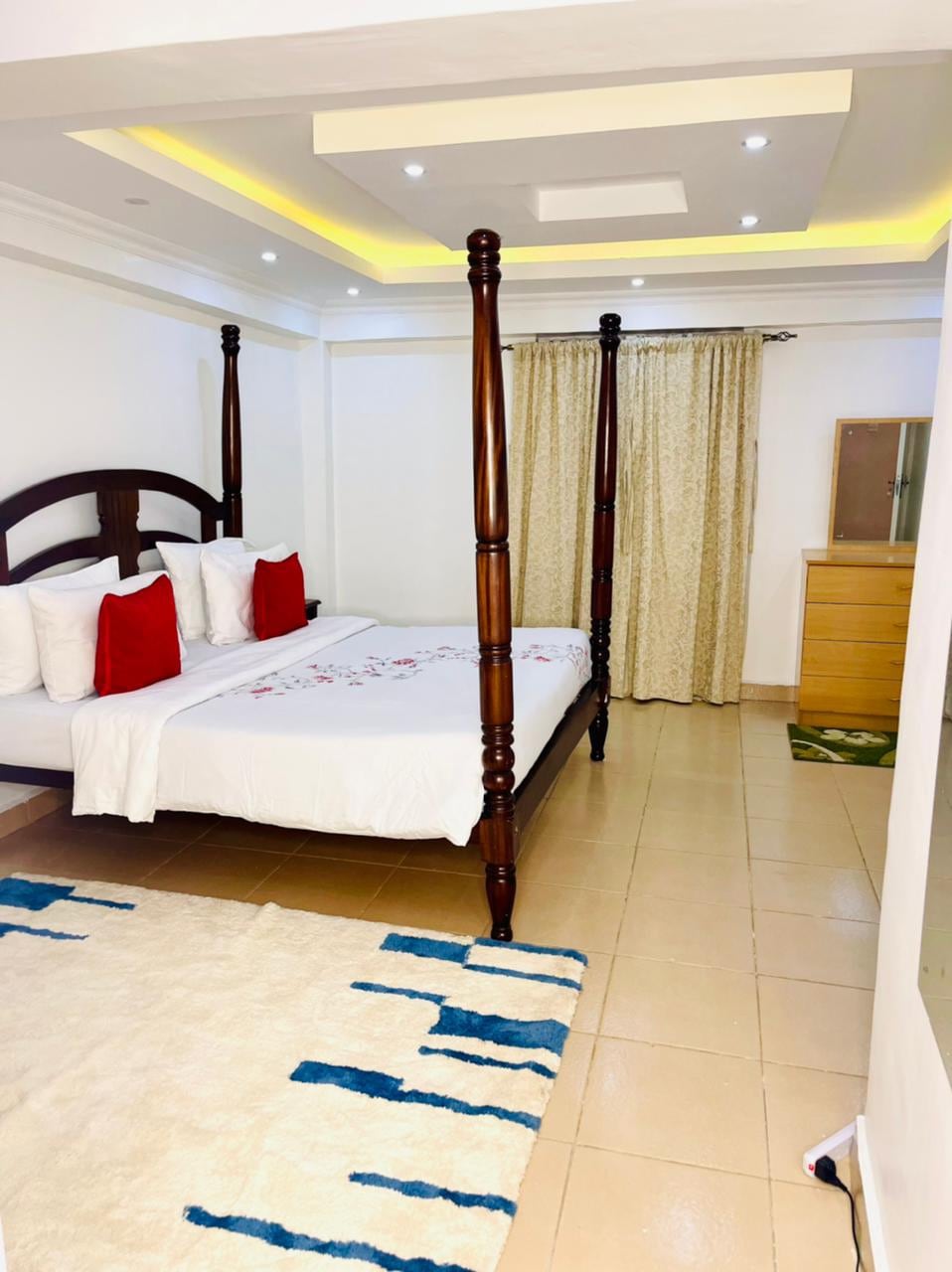 A bright and spacious bedroom features a four-poster bed adorned with white linens and decorative red cushions. A large dresser is positioned nearby, and the room is complemented by a geometric rug that adds a touch of color. Natural light filters through elegant curtains.