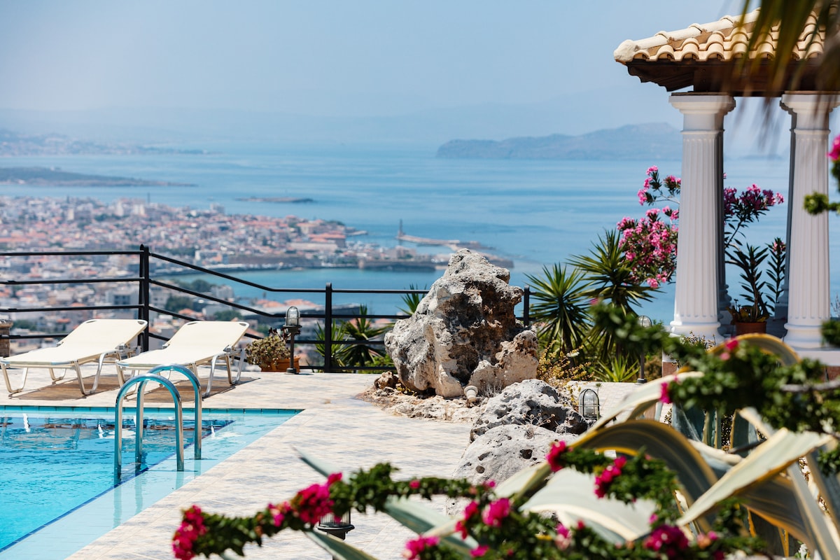 The image captures a stunning view from the patio, featuring a large swimming pool with a sleek railing. Sunbeds are positioned nearby, and lush greenery frames the scene. In the background, the picturesque city of Chania stretches towards the shimmering sea, accentuated by blooming flowers.