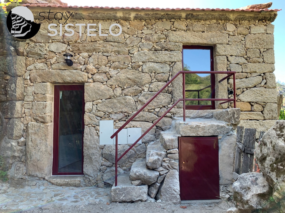 A rustic stone exterior is featured, showcasing textured walls and a red staircase leading to the entrance. Large windows allow natural light into the space, while a wooden door and a small storage unit are visible beside the main entrance.