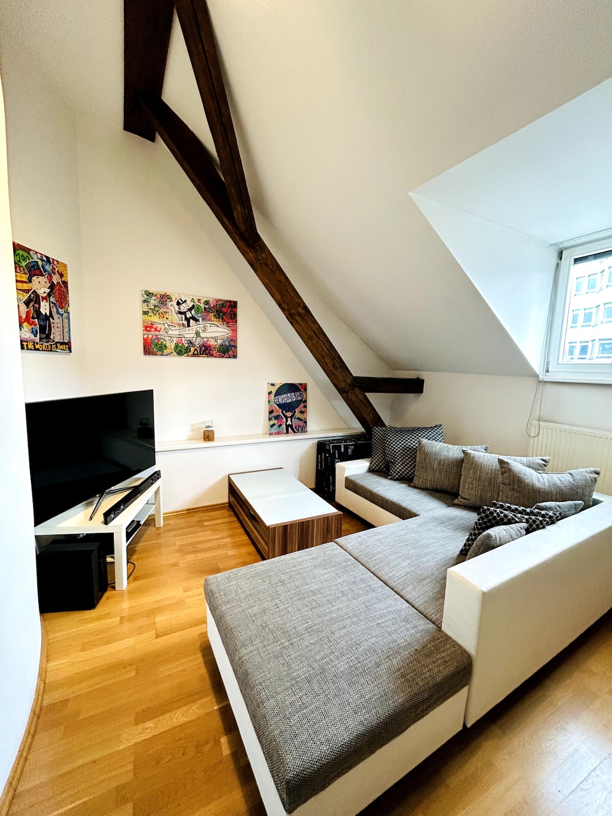 A cozy living area features a plush sectional sofa with soft cushions, positioned near a wall-mounted television. Colorful artwork decorates the walls, while natural light enters through a window with a simple treatment. Wooden beams add character to the lofted space.