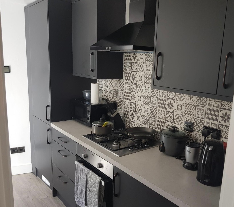 A modern kitchen features sleek black cabinetry and a patterned backsplash. Stainless steel appliances, including a stovetop and microwave, are integrated into the design. Counter space is organized with utensils, a kettle, and cooking tools, ensuring a functional cooking environment.