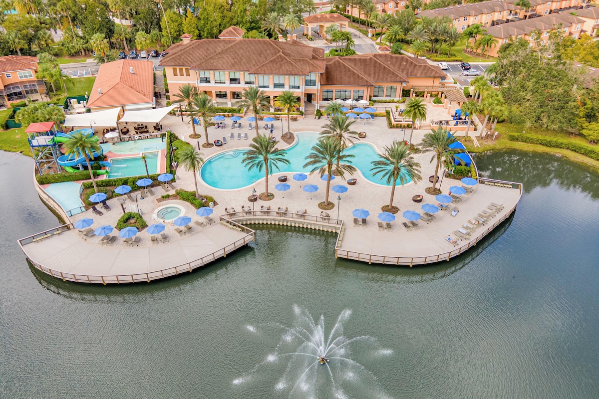 An aerial view of the resort features a large pool area surrounded by palm trees and numerous lounge chairs. A hot tub and a smaller pool are present near the background. The tranquil pond reflects the clear sky, complemented by a fountain at the water's edge.