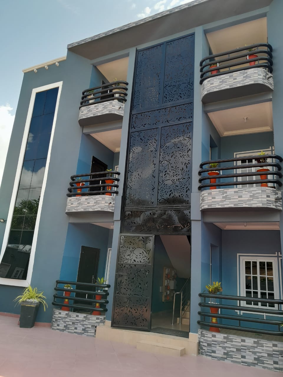 The building features a modern design with a decorative, patterned facade. Large glass windows reflect natural light. Balconies with black railings and potted plants provide outdoor spaces, while the entrance is framed by a stylish stairway leading inside.