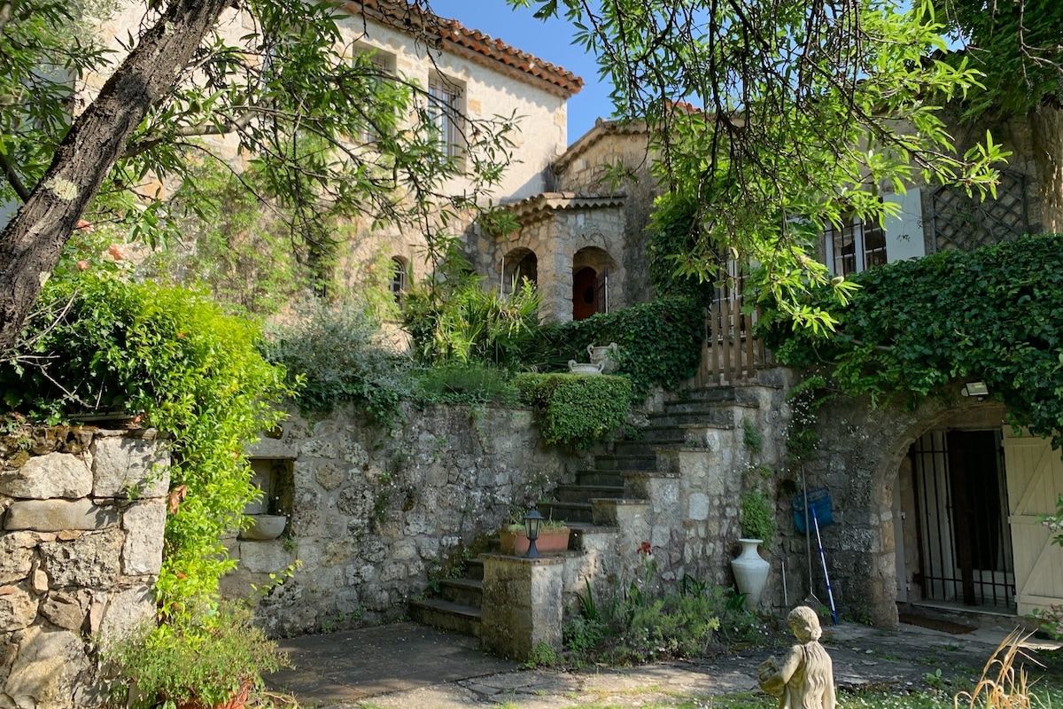 The villa's exterior is framed by lush greenery and rustic stone walls. Stairs lead to the main entrance, surrounded by vibrant plants and sculptures. A sense of warmth is fostered by the sun-drenched walls and shaded areas, inviting peaceful moments outdoors.