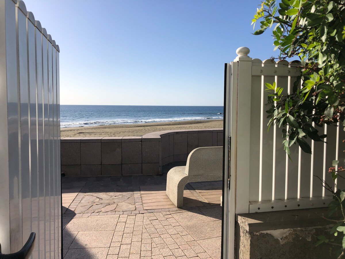 A gate opens to reveal a serene view of a sandy beach and calm ocean waters. A smooth stone bench is positioned on the patio, with sunlight illuminating the space. The scene is framed by a white fence and greenery, enhancing the coastal ambiance.