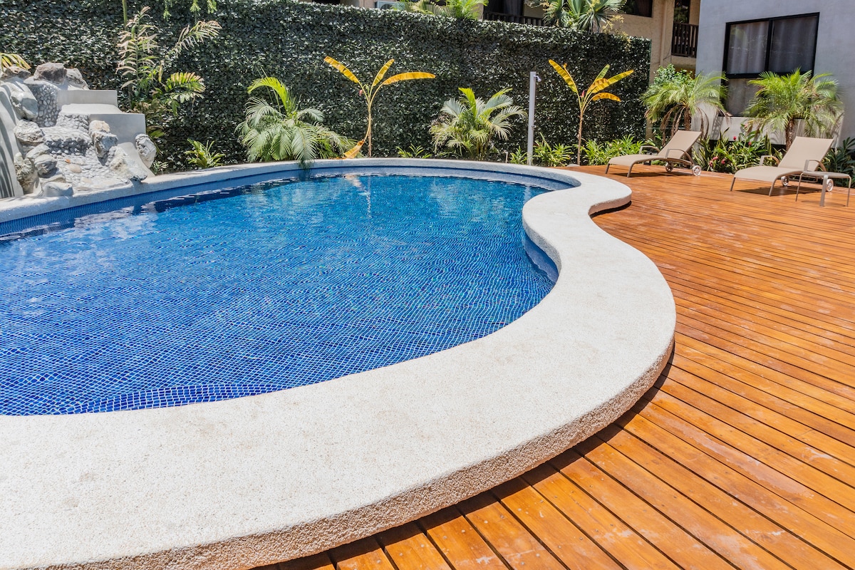 A curvaceous pool features clear blue water, surrounded by a smooth concrete edge. The adjacent wooden deck includes several lounge chairs, and lush green plants provide a tropical ambiance throughout the space.