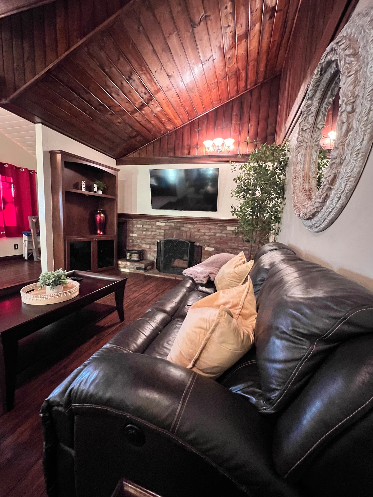 A cozy living area features a dark leather sofa with decorative pillows, positioned near a brick fireplace. A wooden coffee table sits in front, complemented by a wall-mounted television and a large round mirror. Warm lighting enhances the inviting atmosphere.