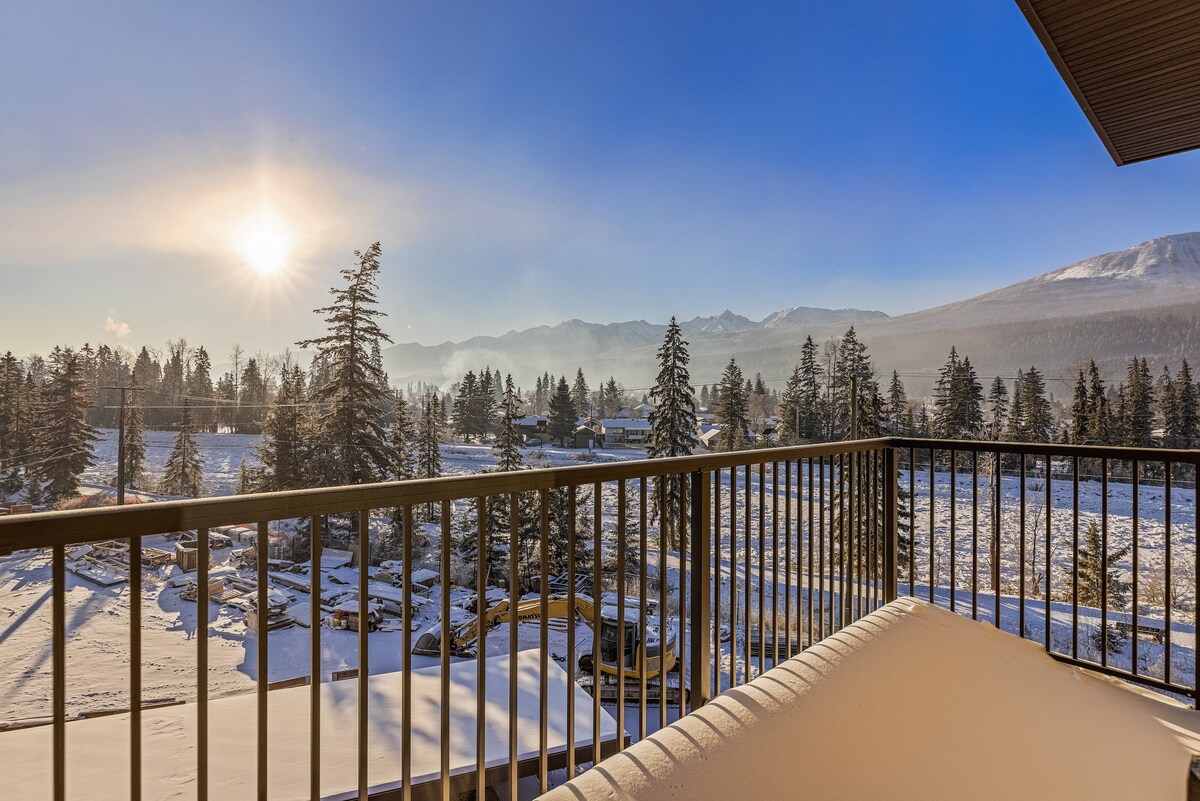 A private balcony offers expansive views of a snow-covered landscape during sunrise. Pine trees frame the scene, with distant mountains visible under a clear blue sky. The tranquil setting is perfect for enjoying the beauty of nature.