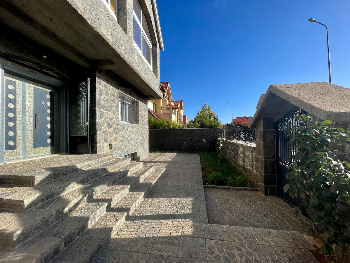 The entrance to the villa features a stone pathway with several steps leading to a large door. The area is surrounded by well-maintained greenery, providing a sense of privacy and calm. Clear blue skies overhead enhance the inviting atmosphere.