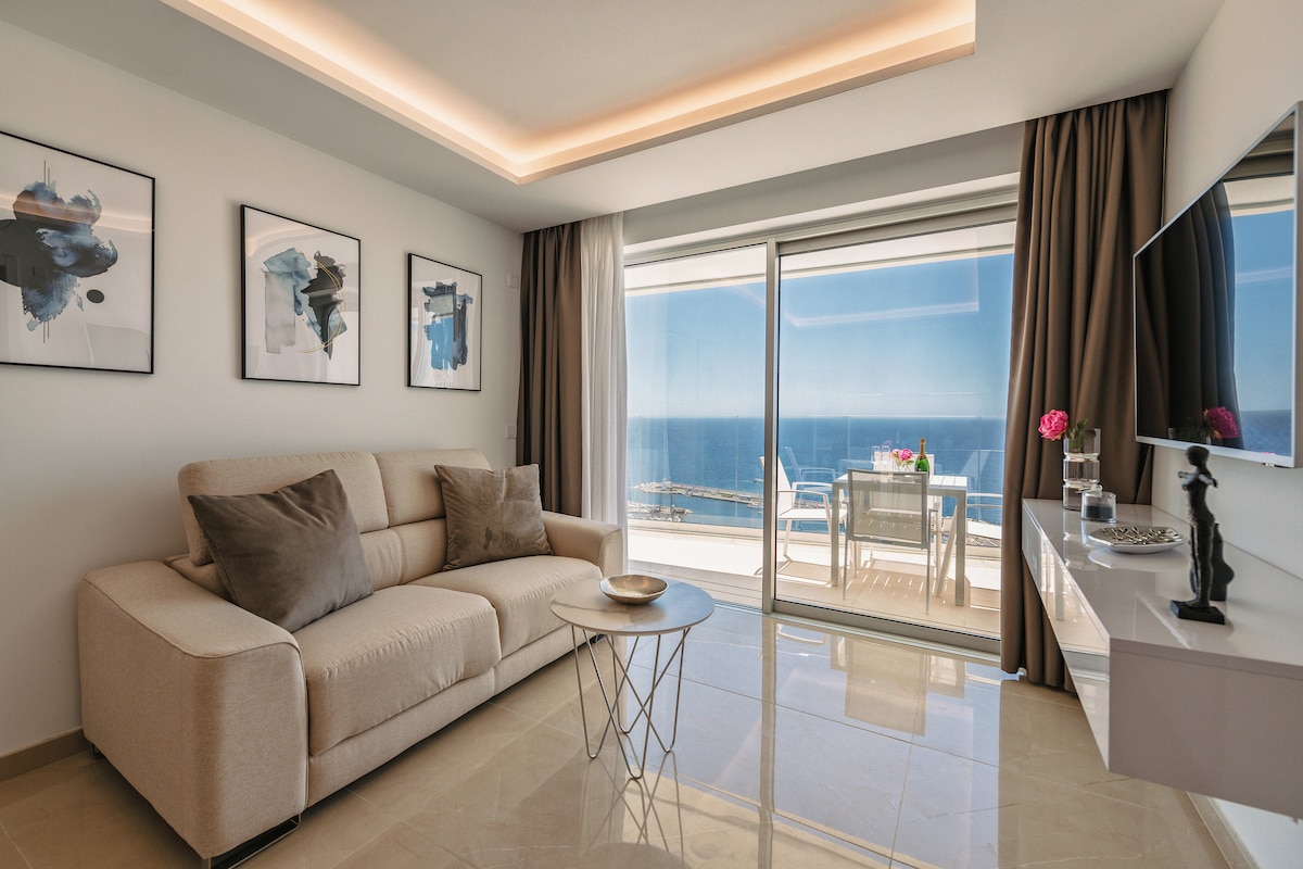 A light-filled living area features a comfortable beige sofa adorned with plush cushions. Two abstract artworks are displayed on the wall, while a sleek flat-screen TV is mounted above a modern console. Floor-to-ceiling glass doors open to a terrace overlooking the ocean.