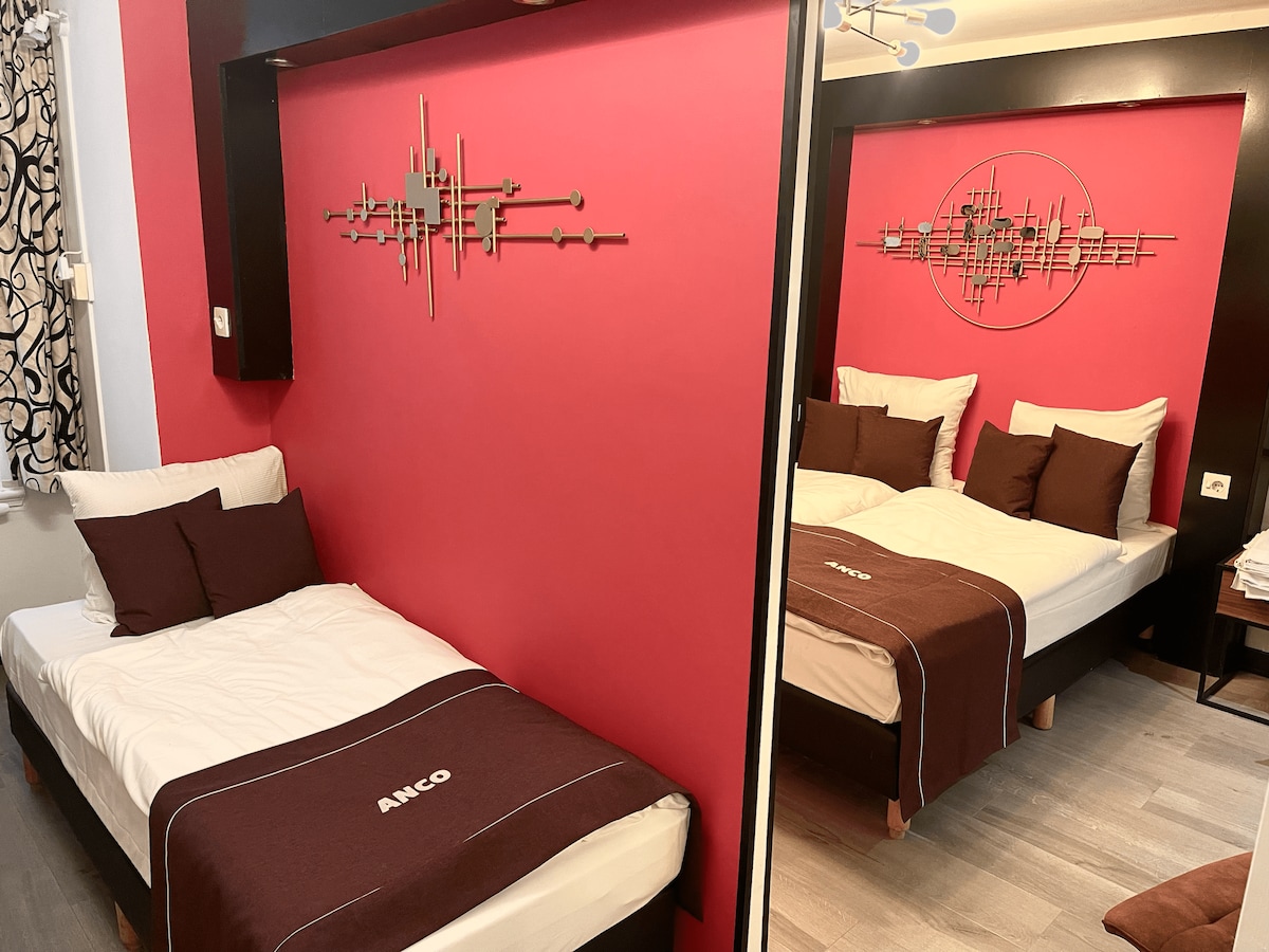 A sleeping area features two single beds with light bedding and dark brown accent pillows. A bold red wall serves as a backdrop, adorned with decorative wall art. A stylish mirror reflects part of the space, enhancing the room's sense of depth.