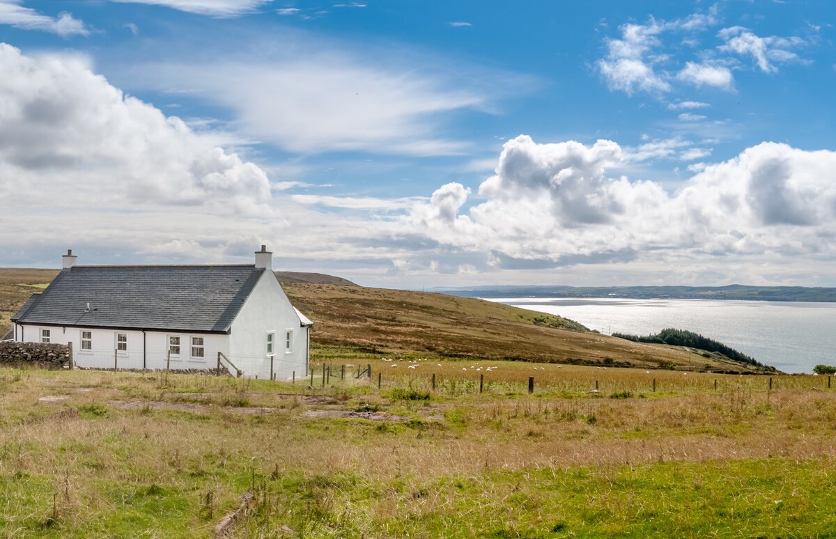A white cottage with a gray roof is situated in an expansive grassy field. Rolling hills are visible in the background, leading to the shimmering waters of Loch Ryan under a bright blue sky adorned with scattered clouds.