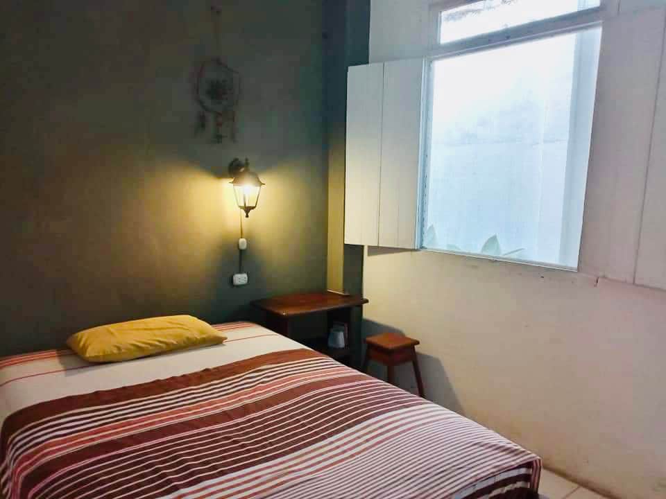 The room features a spacious bed with a striped blanket and a yellow pillow. A small desk and chair are located next to the bed. Natural light enters through the window, which is partially covered by white shutters.