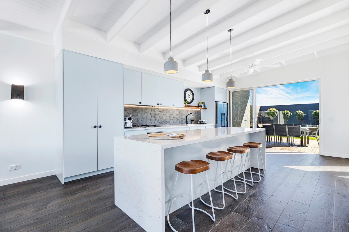 A modern kitchen features a large island with four wooden stools, complemented by light grey cabinetry and stylish pendant lights. A spacious living area is visible through sliding glass doors, revealing an outdoor dining space and inviting natural light.