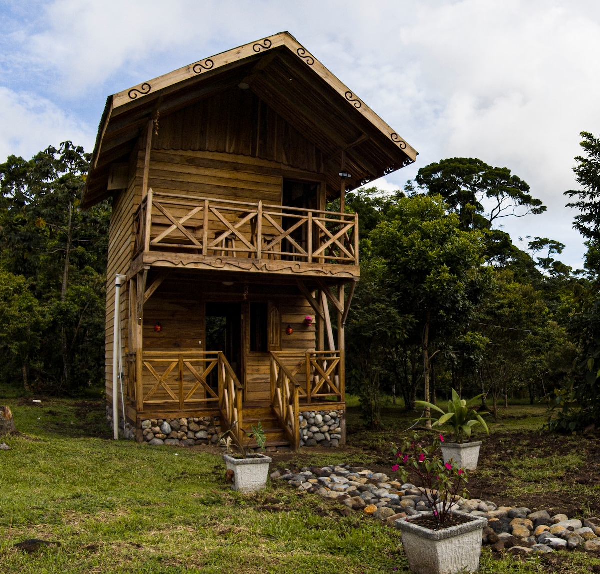 A wooden cottage stands surrounded by lush greenery, featuring a spacious balcony with intricate railings. The building is elevated on stone pillars, and the entrance is accentuated by a flight of stairs, inviting guests to explore the natural surroundings.