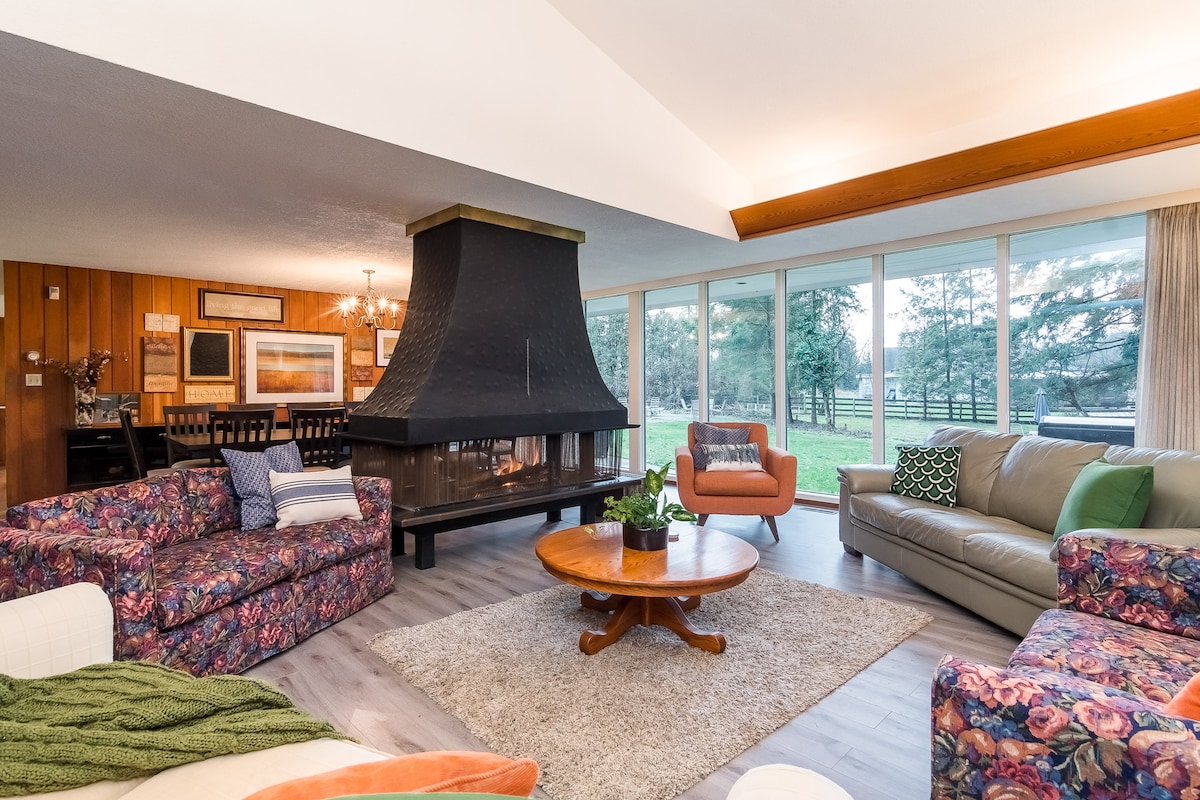 A spacious living area features colorful seating options, including floral-patterned and neutral-toned sofas. A central wood-burning fireplace adds warmth, while large windows allow natural light to illuminate the space and offer views of the surrounding greenery.