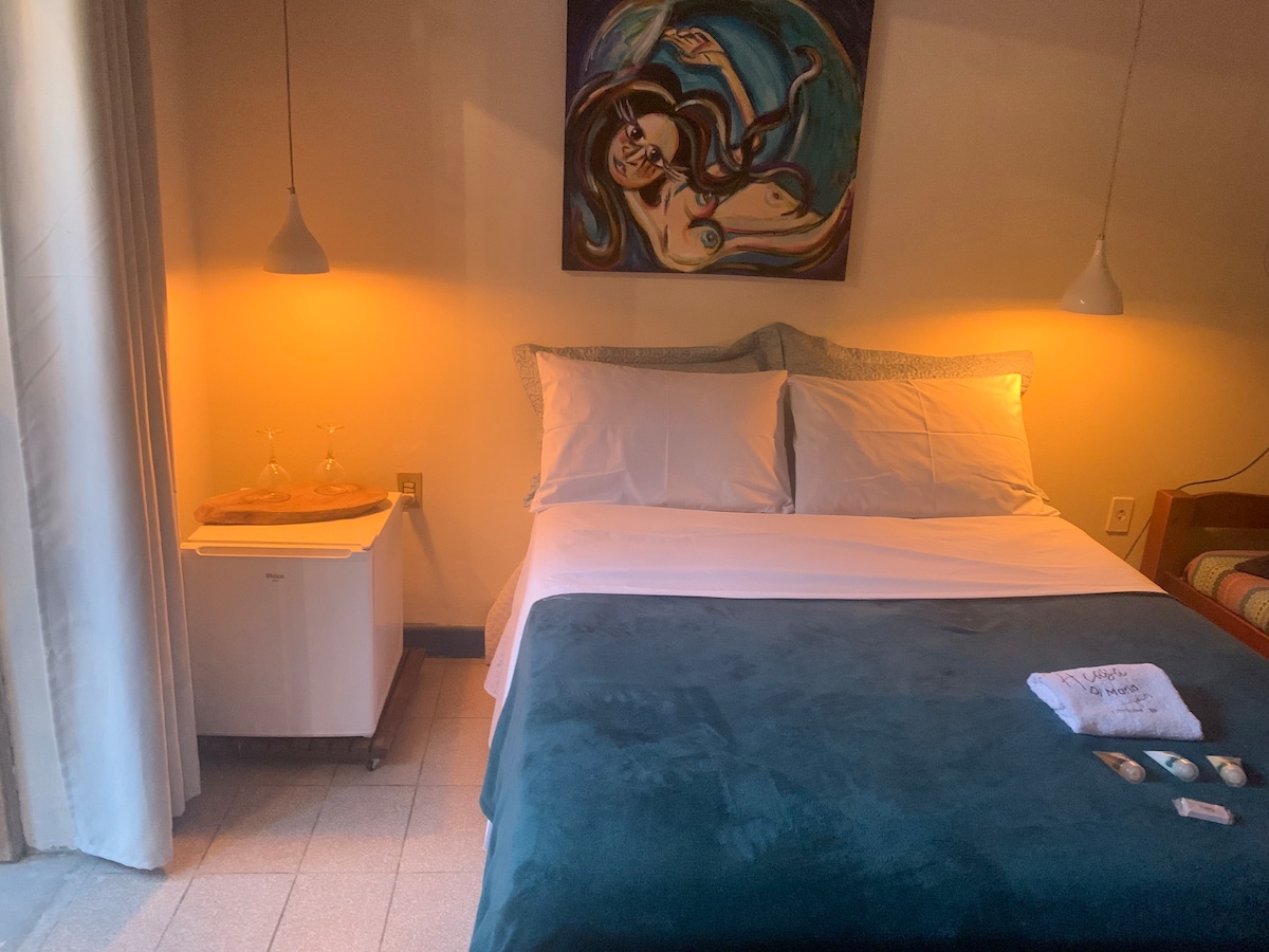 A comfortable sleeping area features a spacious bed layered with soft linens and a dark blanket. Subtle lighting creates a warm ambiance. A piece of wall art adds interest above the bed, while a small side table holds clear glasses. A compact refrigerator is positioned beside the bed.