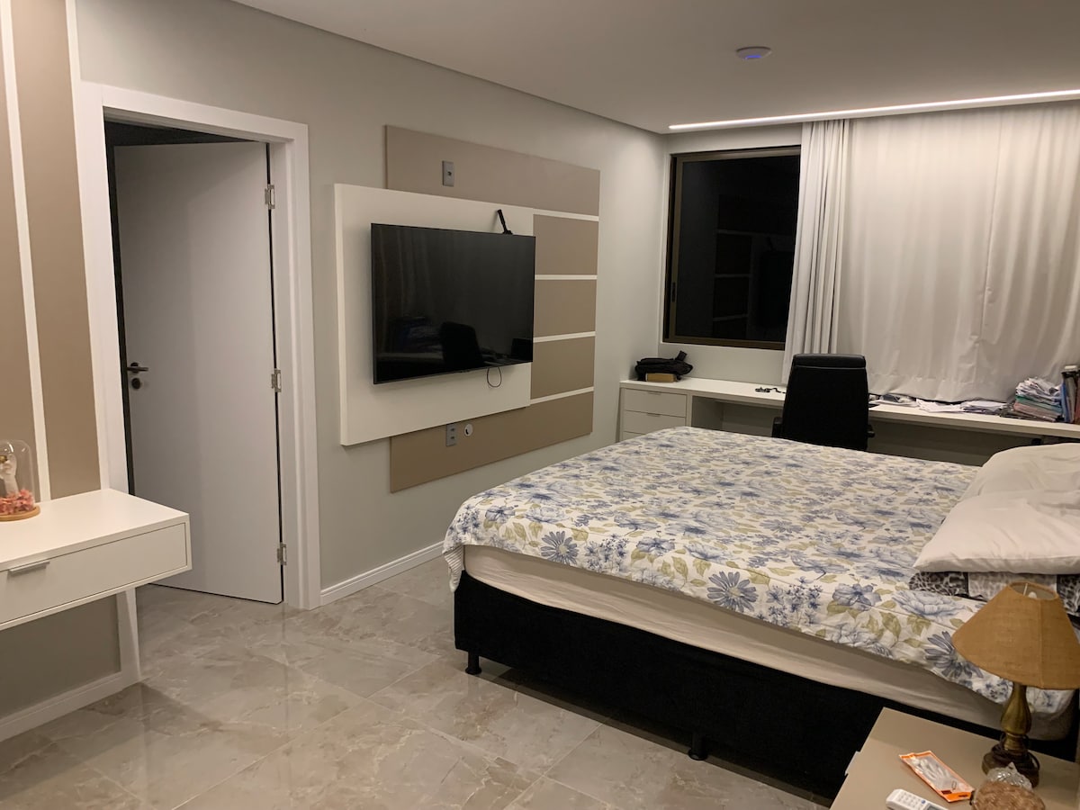 A spacious bedroom is presented, featuring a large bed with a floral-patterned blanket. A television is mounted on the wall opposite the bed. Natural light filters through a large window adorned with light curtains, and a modern desk chair is positioned beside a neatly organized workspace.