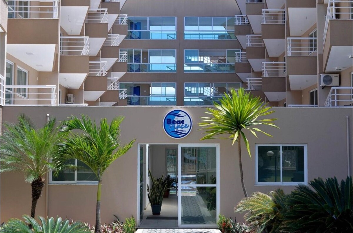 The entrance of Beach Place Cumbuco is depicted, showcasing a modern building with a clear sign displaying the property name. Lush palm trees frame the entrance, while large windows on the upper floors reflect natural light, suggesting a welcoming environment.
