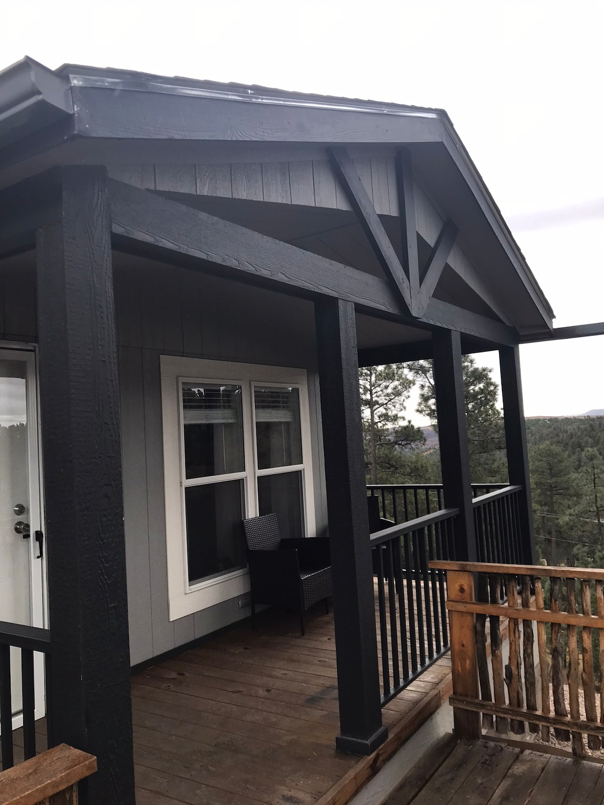 A covered porch area features wooden decking and black railing, providing an outdoor seating space. Two chairs are positioned to enjoy serene views of the surrounding trees and hills. Large windows are visible behind the porch, allowing natural light into the interior.