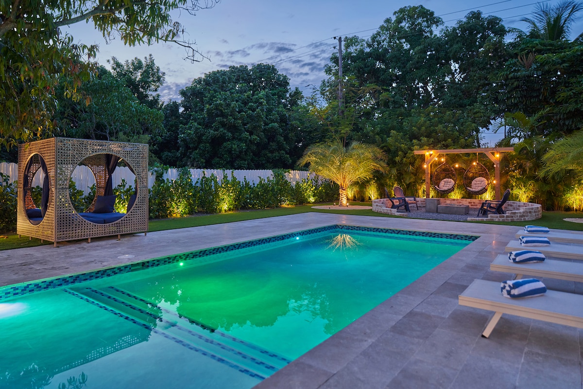 The outdoor area features a heated pool illuminated with soft lighting, surrounded by well-maintained greenery. A cozy seating area with a fire pit is present nearby, along with two circular hanging chairs for relaxation, creating an inviting space for outdoor enjoyment.