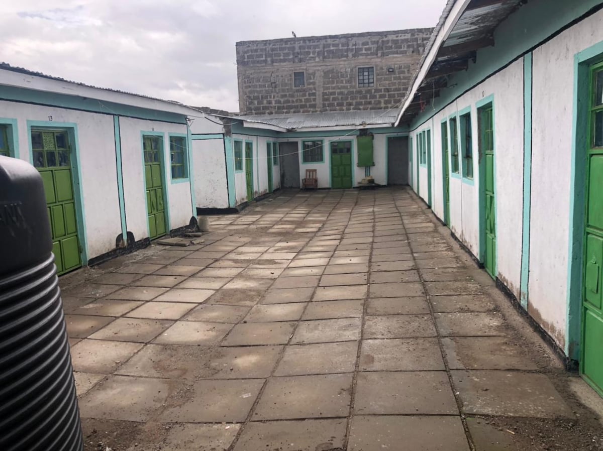 An enclosed courtyard is showcased, featuring a stone-paved surface. Green doors line the walls of the surrounding buildings, providing access to rooms. A few wooden chairs are visible in the background, and the sky appears overcast, contributing to a calm atmosphere.