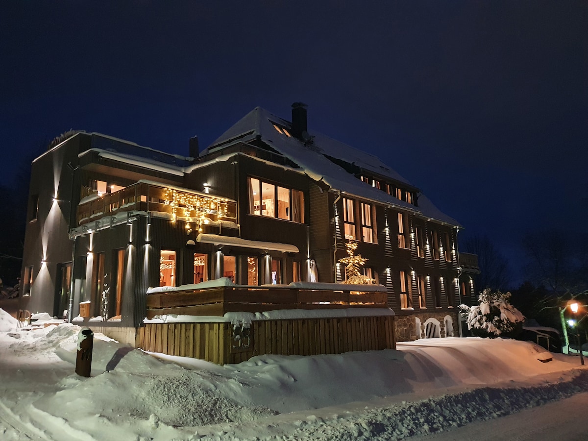 The lodge is illuminated by soft lighting against a winter night backdrop. Fresh snow blankets the surroundings, while large windows reveal warm indoor spaces. The exterior features a spacious deck and elegant architectural details, showcasing the alpine style of the accommodation.