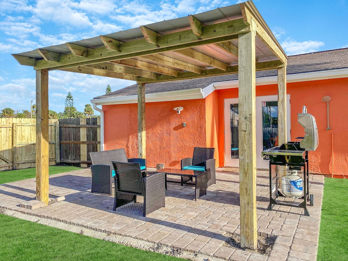 An outdoor patio features a wooden pergola providing shade over a seating area with dark wicker furniture and teal cushions. A grill is positioned nearby for outdoor cooking. The vibrant orange wall of the house adds a pop of color, while the lawn is neatly trimmed.