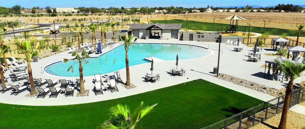 A spacious community pool is surrounded by palm trees and lounge chairs, offering ample seating. A shaded structure provides a gathering area, while a grassy area adds to the outdoor appeal. The distant landscape features gently rolling terrain, creating a relaxed atmosphere for guests.