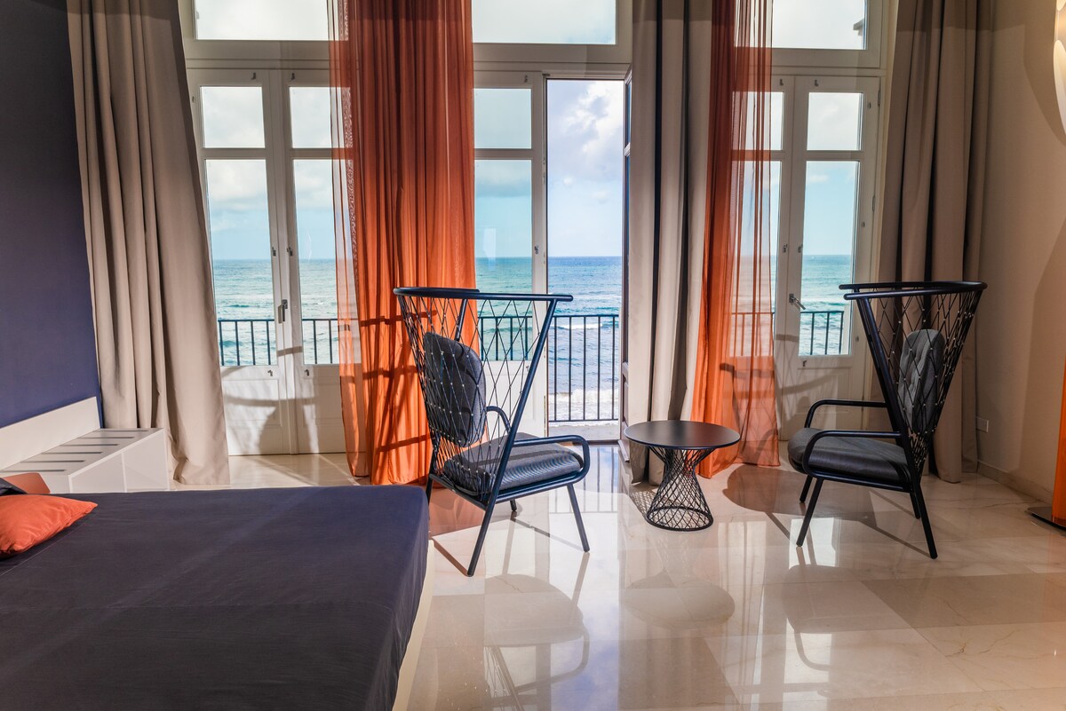 The room features large windows adorned with flowing curtains, offering views of the sea. Two stylish chairs are positioned around a small table on the polished floor, alongside a spacious bed with neutral bedding, creating a blend of comfort and elegance.