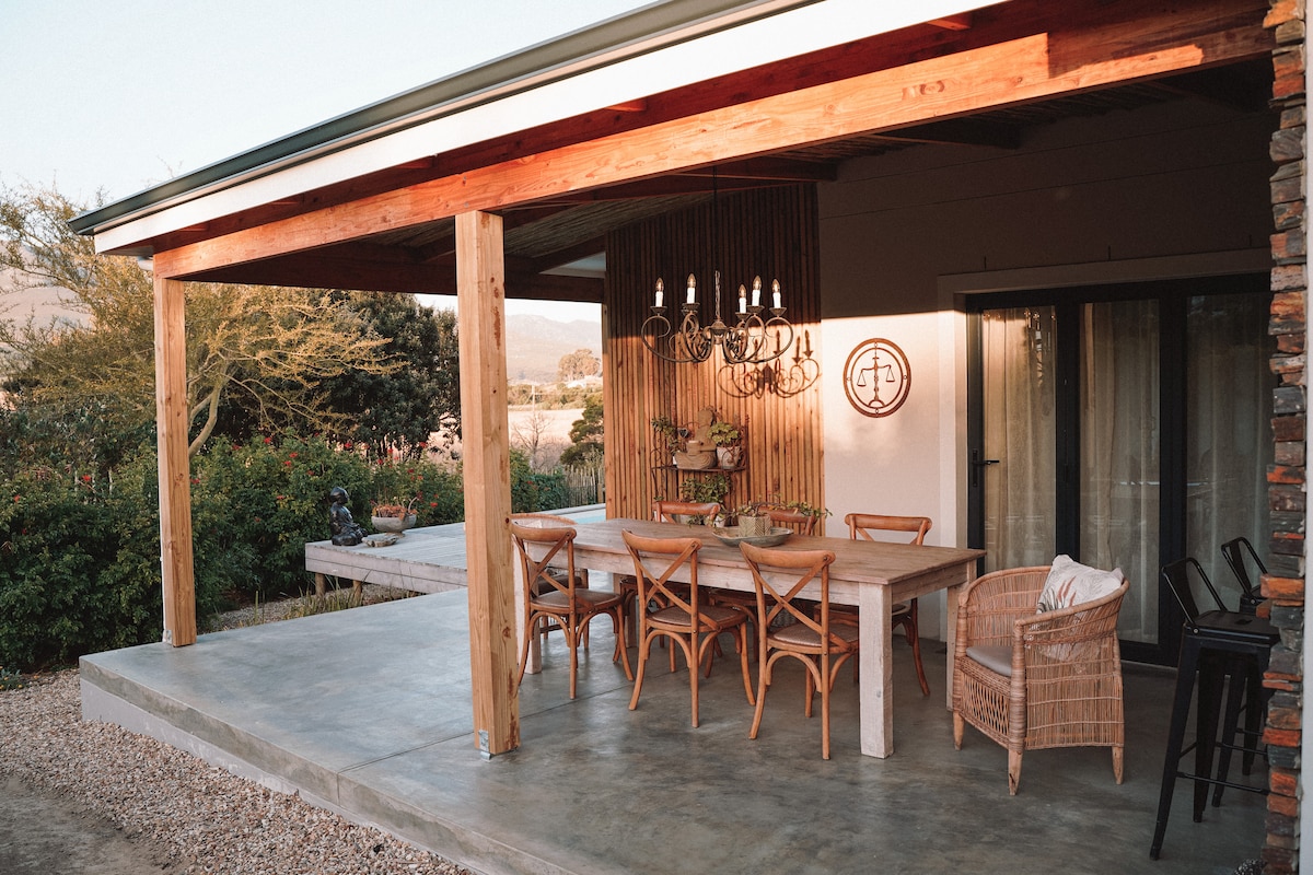 An outdoor dining area is presented under a wooden pergola, featuring a long, rustic table surrounded by mixed seating options, including wicker chairs. Natural light casts gentle shadows, enhancing the surrounding greenery and creating a serene outdoor atmosphere.
